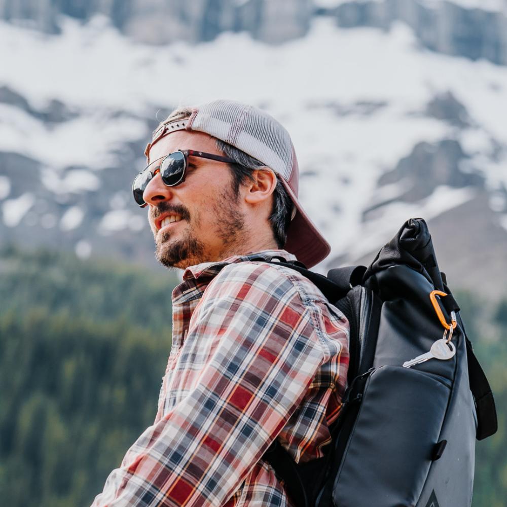 Homme avec sac à dos sur fond de sapins et montagne portant des lunettes équipées de clip on solaires